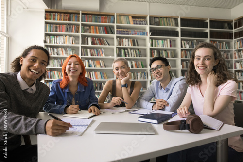 Happy diverse college fresh students posing for group portrait in campus library, sitting at table with bookshelves in background, writing notes, studying books, looking at camera, smiling