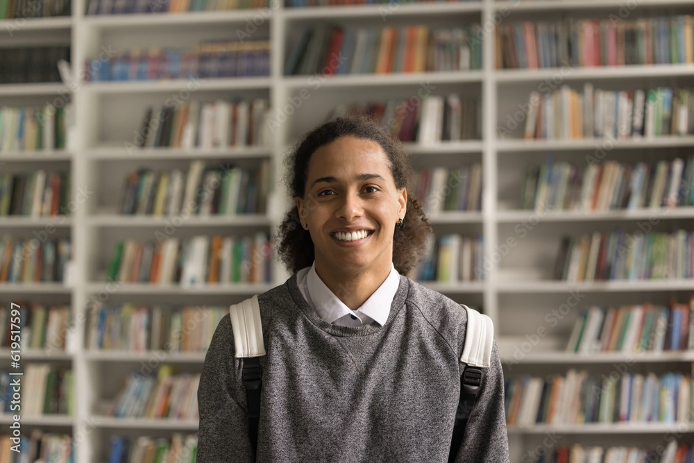 Cheerful long haired fresh African American student guy with backpack standing in campus, university library with bookshelves in background, looking at camera with toothy smile. Head shot portrait Cheerful long haired fresh African American student guy with backpack standing in campus, university library with bookshelves in background, looking at camera with toothy smile. Head shot portrait