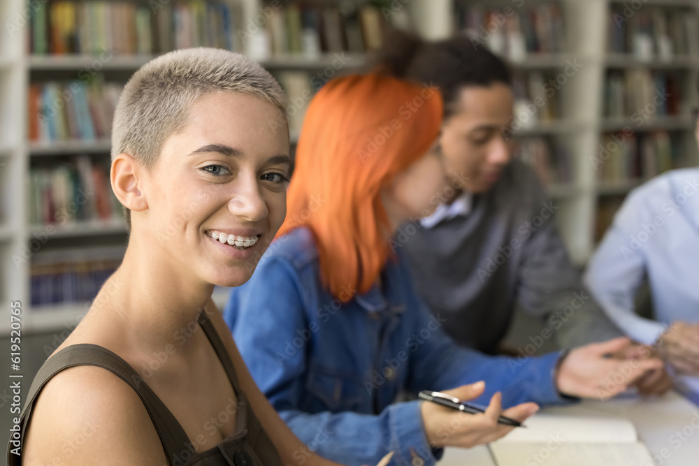 Happy trendy young student girl studying in university library, sitting at laptop, looking at camera, posing, sitting at table with diverse classmates in background, smiling, showing teeth brackets Happy trendy young student girl studying in university library, sitting at laptop, looking at camera, posing, sitting at table with diverse classmates in background, smiling, showing teeth brackets