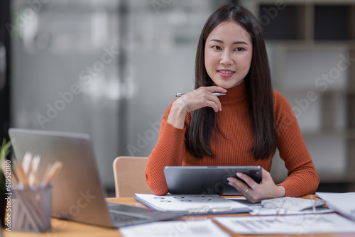 Attractive smiling young asian business woman wear glasses work at home office, Asian woman working on laptop computer holding tablet.business finance concept.