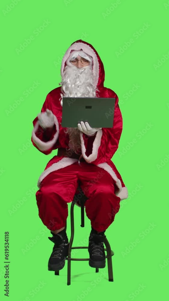 Vertical video Front view of modern santa character on laptop in full body greenscreen studio, sitting on chair and browsing social media app on wireless pc. Young cool saint nick cosplay in red Vertical video Front view of modern santa character on laptop in full body greenscreen studio, sitting on chair and browsing social media app on wireless pc. Young cool saint nick cosplay in red