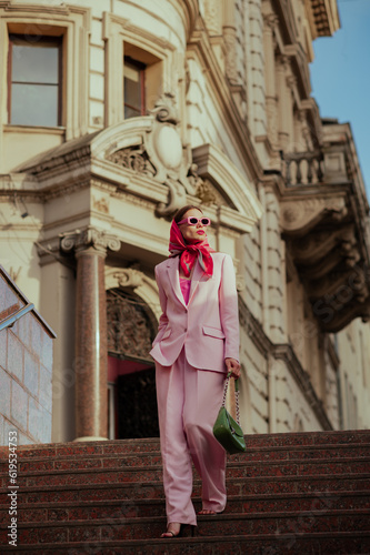 Fashionable elegant confident woman wearing trendy pink sunglasses, silk headscarf, suit blazer, wide leg trousers, with green bag, walking in street of European city. Outdoor full-length portrait