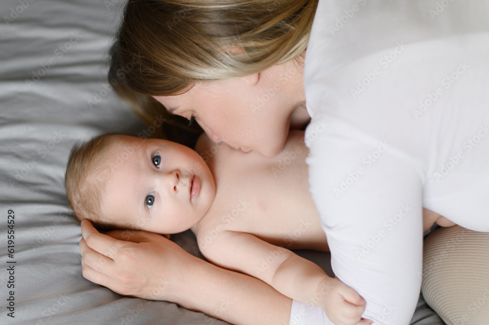 Joyful young mother plays with her baby lying on the bed, smiles, kisses the cheek, enjoys motherhood.Family, love,care. Joyful young mother plays with her baby lying on the bed, smiles, kisses the cheek, enjoys motherhood.Family, love,care.