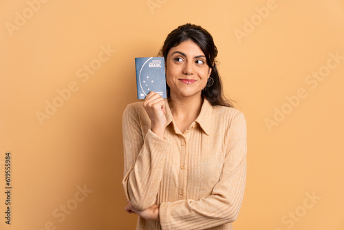 cheerful brazilian woman showing brazilian passport in beige studio background. travel, trip, brazil concept.