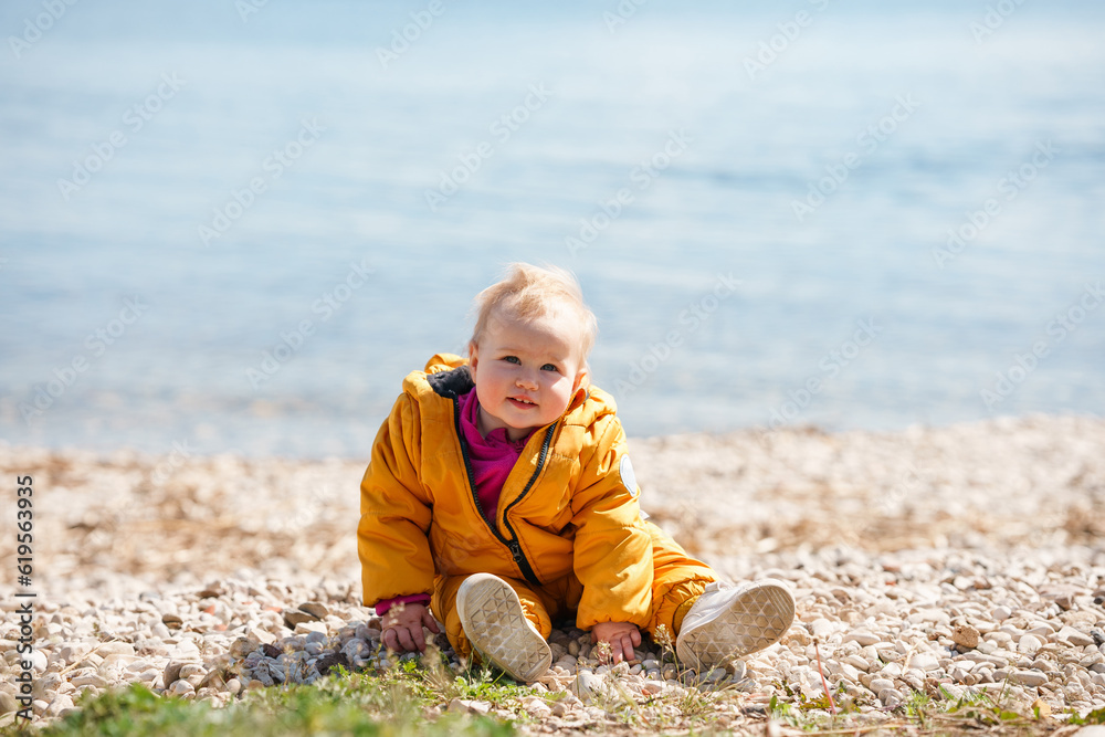 Cute baby toddler sitting on pebbles by the sea in the cold season Cute baby toddler sitting on pebbles by the sea in the cold season
