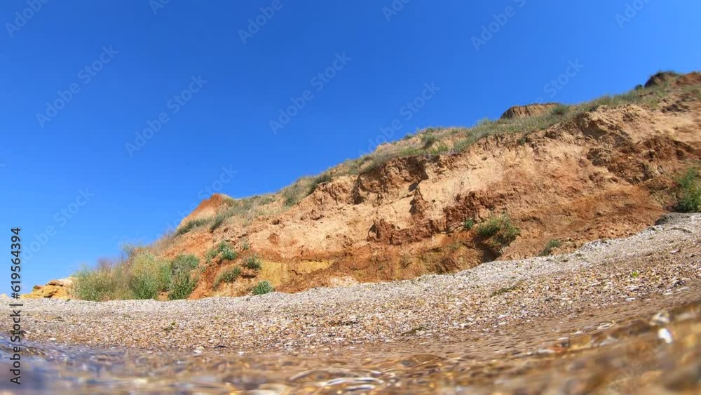 View of coast with mountain and blue sky from under below transparent water on sandy beach of sea coast. Sunny summer day. Wide below angle. Moving sea wave inside. Nature background. Ripples on water View of coast with mountain and blue sky from under below transparent water on sandy beach of sea coast. Sunny summer day. Wide below angle. Moving sea wave inside. Nature background. Ripples on water