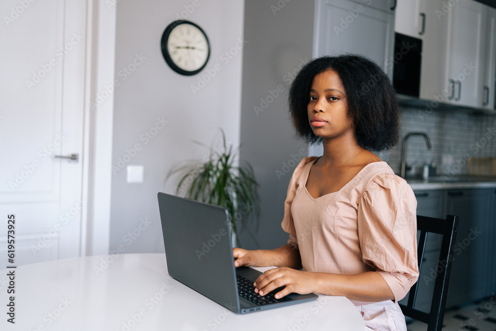 Fototapeta premium African American young business woman working or studying remotely from home office using laptop siting at desk in modern kitchen room, serious looking at camera. Black young woman doing online job.