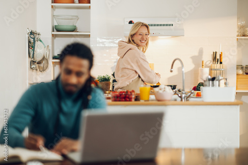 A happy housewife is cooking in a kitchen while her Arabic husband is working in a blurry foreground.