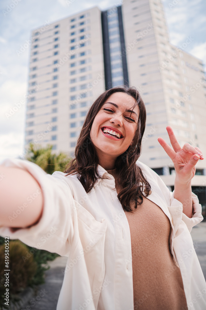 Vertical portrait of pretty young adult woman with thooty smile having fun taking a selfie using a social media. Happy teenage female laughing and looking at camera with cheerful expression outside