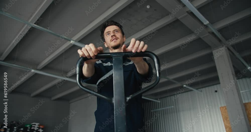 Close-up shot of a man in a special blue uniform pulling a load by the handle of a dragging machine. Warehouse worker transports and sorts goods Close-up shot of a man in a special blue uniform pulling a load by the handle of a dragging machine. Warehouse worker transports and sorts goods