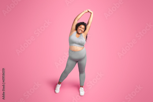 Positive black plus size lady in sportswear doing arm stretching exercises, warming up before training, pink background