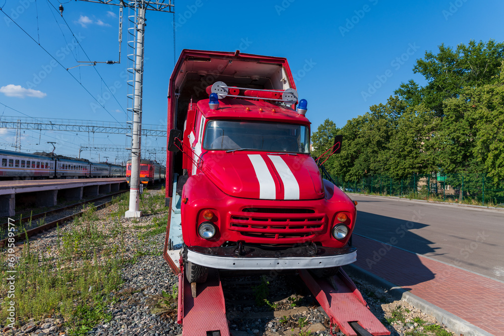 Red fire engine pulls out of freight covered wagon on railway station in a summer sunny day. No people. Clear blue sky. Firefighting apparatus theme.