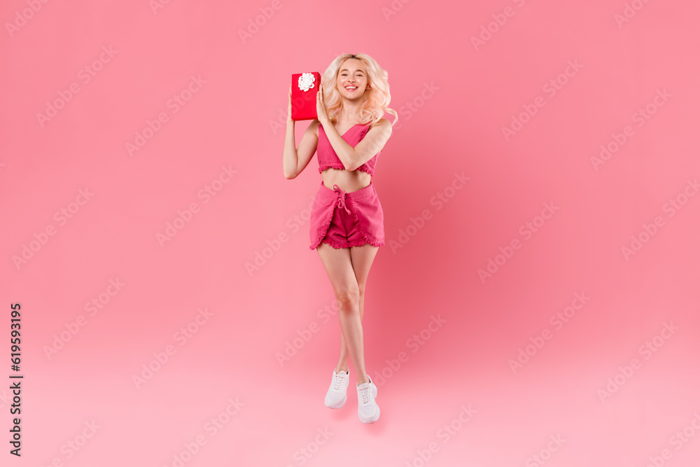 Happy pretty young woman jumping and holding wrapped gift box smiling at camera isolated on pink background, full length Happy pretty young woman jumping and holding wrapped gift box smiling at camera isolated on pink background, full length