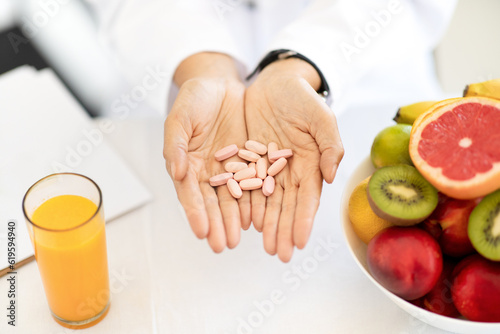 Fototapeta Naklejka Na Ścianę i Meble -  Adult european woman doctor nutritionist in white coat at table with organic fruits and juice holds pills in hands