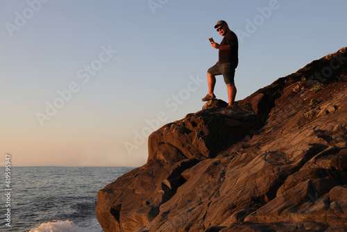 silhouette of tourist man stand of hill mountains at the sunset rays. nature background. male gay looking in screen of mobile cell phone in hand. male backpacker. Batumi  Georgia.