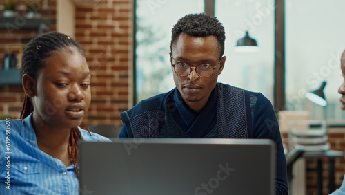 Company team at desk planning reports, reading business statistics on laptop. African american people analyzing corporate results with official information, employees with expertise.