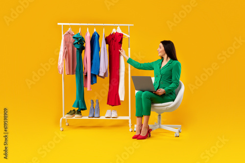 Online consultation with stylist. Happy young woman sitting with laptop computer near clothing rack, yellow background