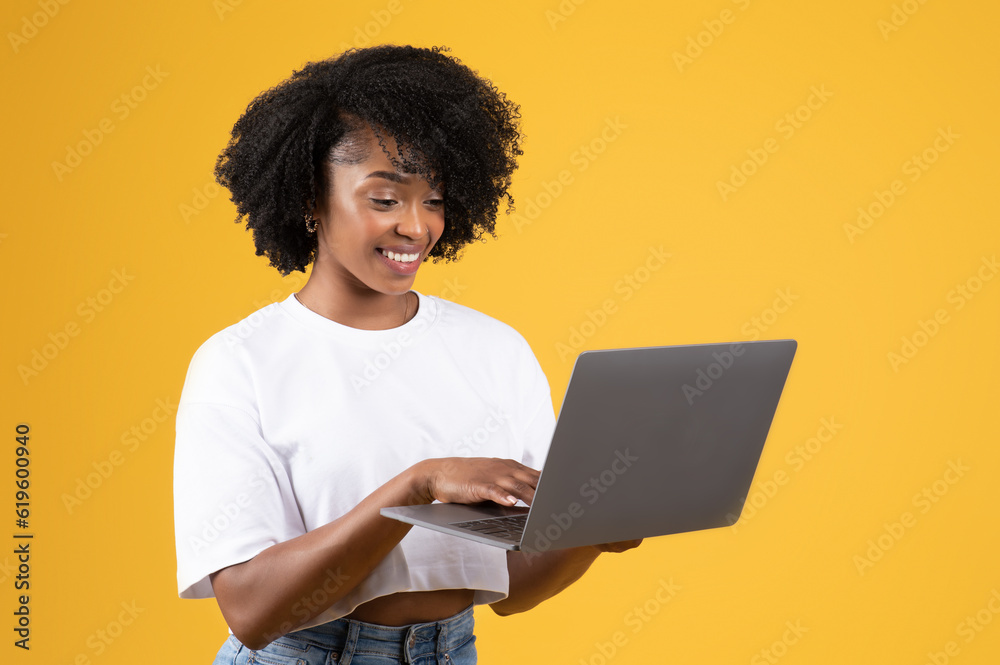 Smiling pretty millennial black lady in casual typing on laptop, isolated on orange studio background Smiling pretty millennial black lady in casual typing on laptop, isolated on orange studio background