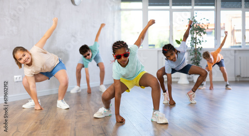 African girlie perform movements during warm-up, limbering-up part of workout with peers. Group of young girls and guys in dark glasses dance in fitness club in background unfocused