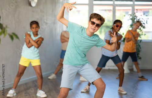 Boy in sun glasses performs choreographic exercises and teaches energetic mobile social dance Jazz-modern together with friends. Multi-racial group of children performs movements in spacious studio
