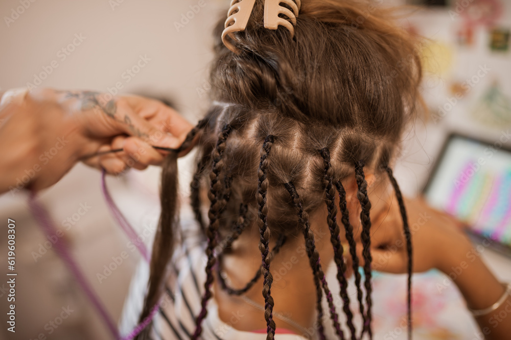 A mother braids her teenage daughter in many thin African braids while she draws on a tablet in her room.