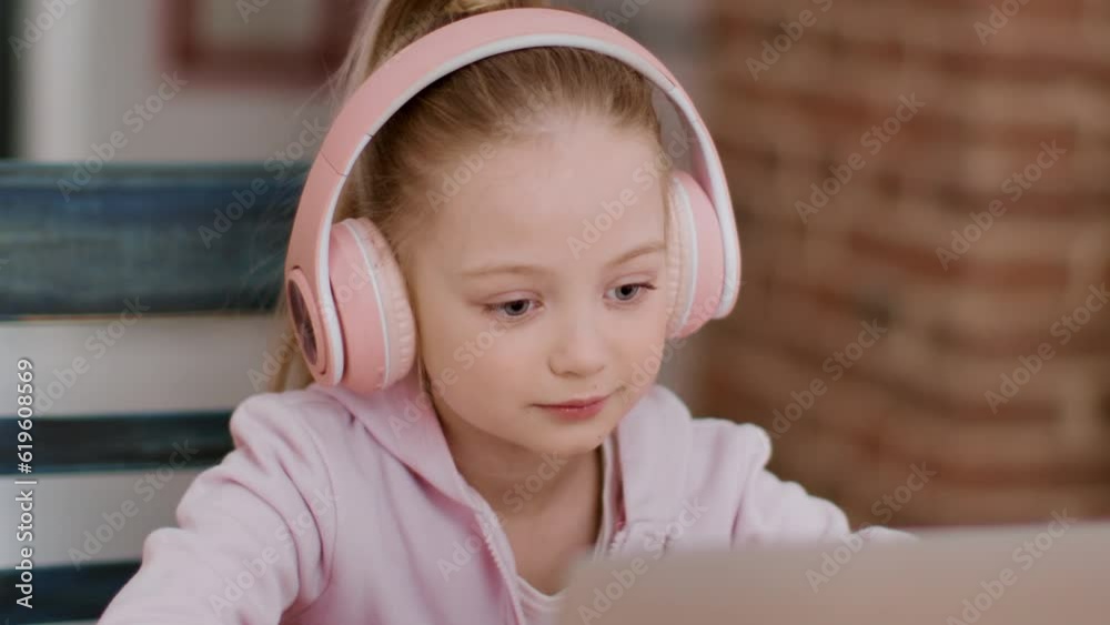 Primary online education. Smart little girl in headphones studying at home, watching lesson on laptop and writing task Primary online education. Smart little girl in headphones studying at home, watching lesson on laptop and writing task