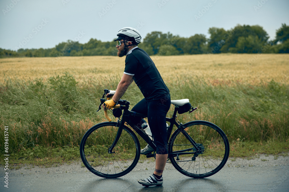 Young bearded man, cyclist in uniform, helmet and glasses standing with bicycle on road around field background. Concept of sport, hobby, leisure activity, training, health, speed, endurance, ad