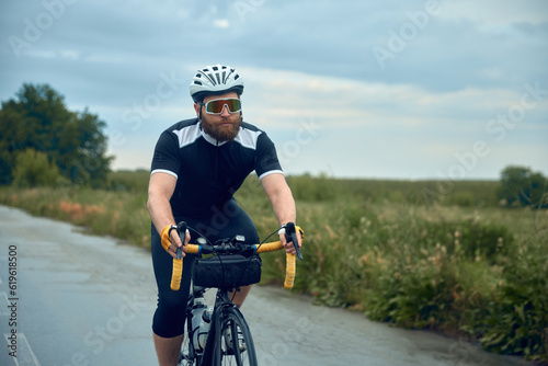 Young bearded man, cyclist in uniform, helmet and glasses ridin bicycle in chill evening after rain. Concept of sport, hobby, leisure activity, training, health, speed, endurance, ad