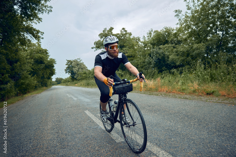 Young bearded man, cyclist in helmet, glasses and uniform riding bike on wet road in cloudy chill evening. Concept of sport, hobby, leisure activity, training, health, speed, endurance, ad Young bearded man, cyclist in helmet, glasses and uniform riding bike on wet road in cloudy chill evening. Concept of sport, hobby, leisure activity, training, health, speed, endurance, ad