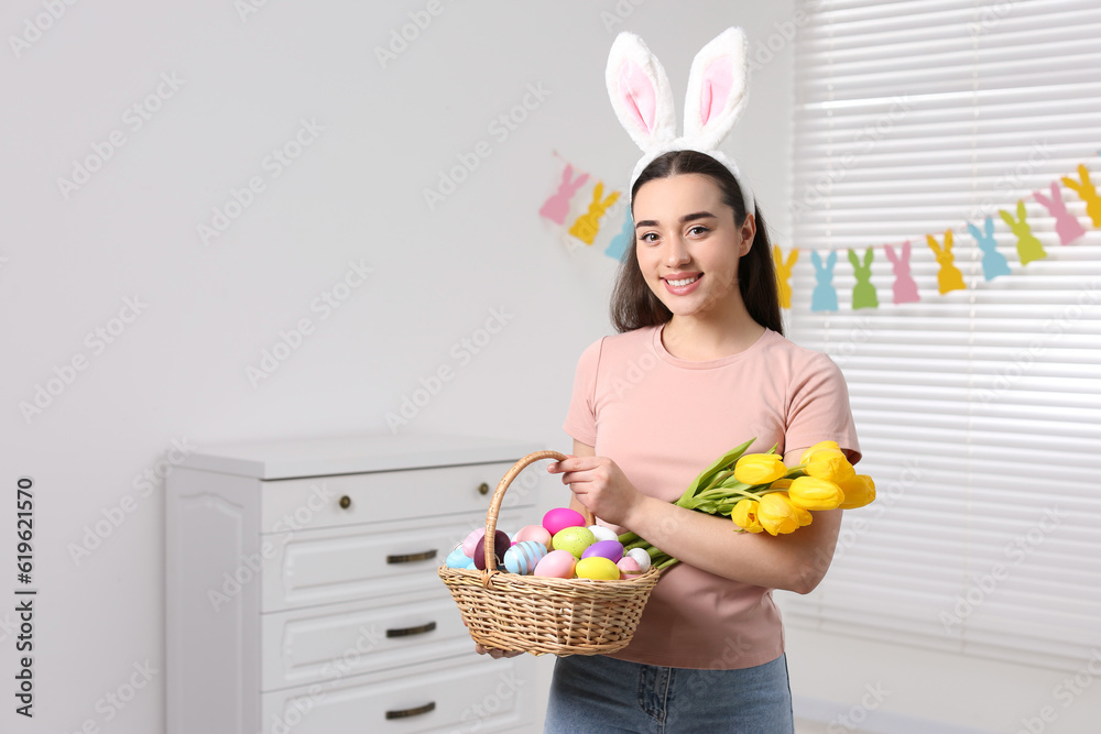 Happy woman in bunny ears headband holding wicker basket with painted Easter eggs and bouquet of flowers indoors. Space for text