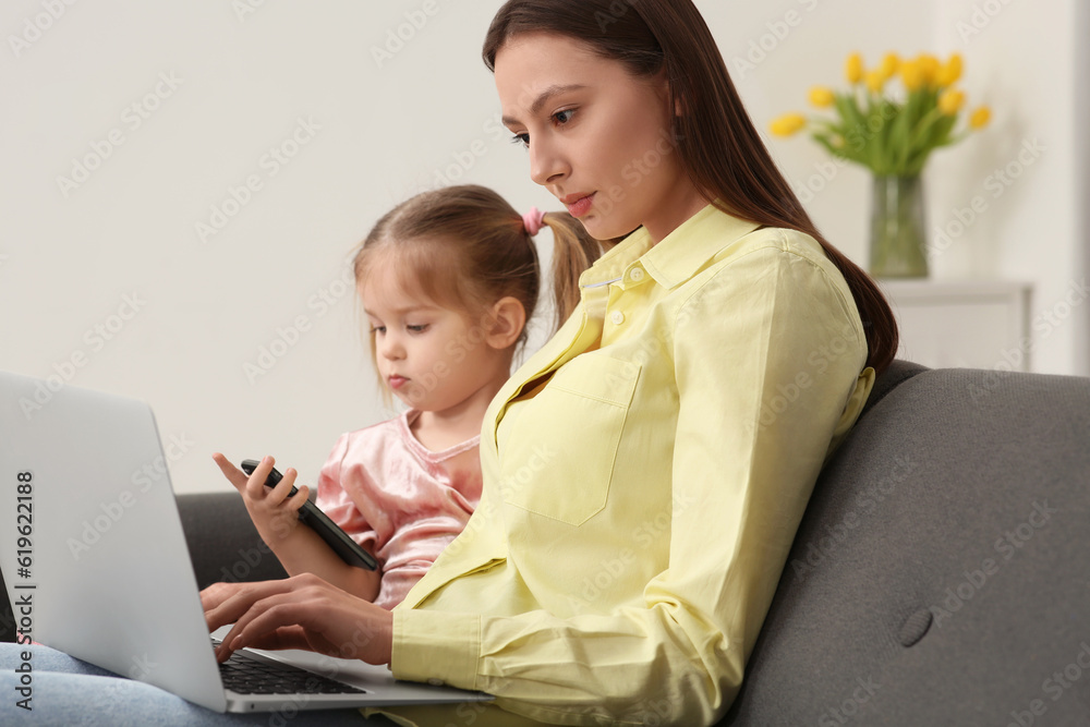 Mother working remotely on laptop while her daughter playing with smartphone at home Mother working remotely on laptop while her daughter playing with smartphone at home