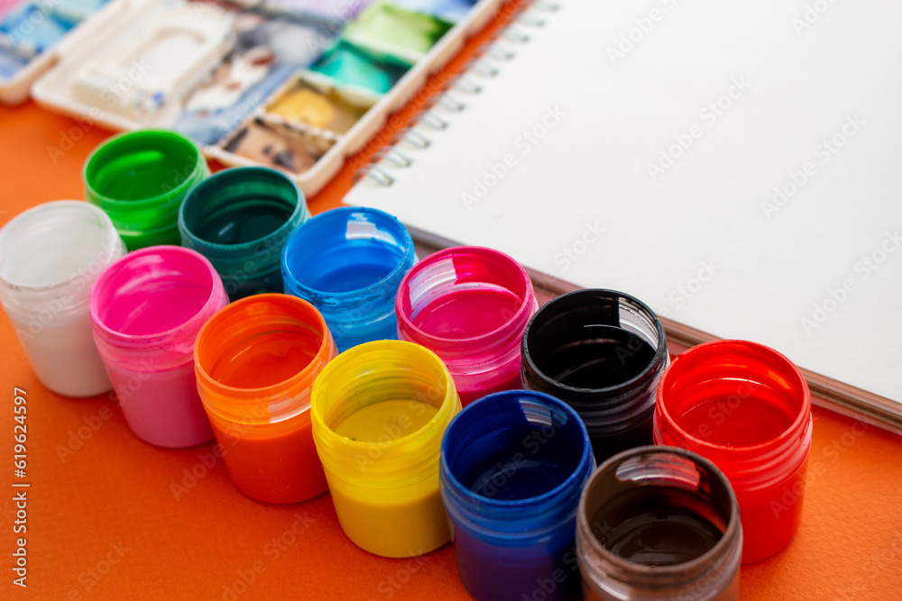 Potes de tinta guache de diversas cores, com caderno e godê ao fundo Potes de tinta guache de diversas cores, com caderno e godê ao fundo