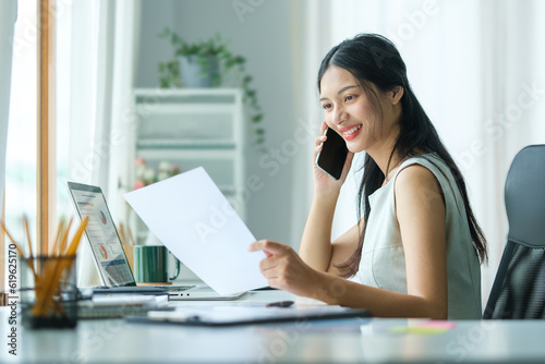 Smiling businesswoman holding document and working with laptop computer in bright office.
