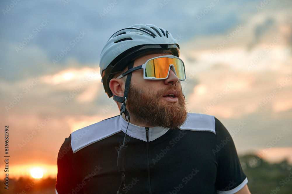 Close-up portrait of young bearded man, cyclist in helmet and special glasses standing over sunset sky background. Concept of sport, hobby, leisure activity, training, health, speed, endurance, ad Close-up portrait of young bearded man, cyclist in helmet and special glasses standing over sunset sky background. Concept of sport, hobby, leisure activity, training, health, speed, endurance, ad