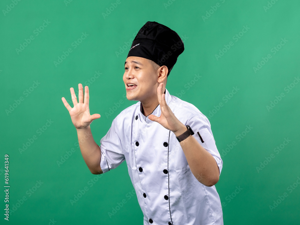 A portrait of an Indonesian Asian man wearing a chef's uniform and a chef's hat, opening his hands and posing as if shouting, isolated with a green background.