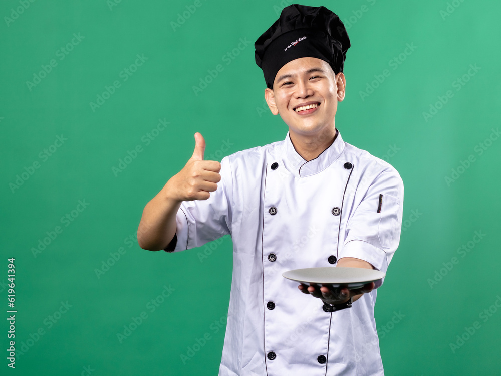 A portrait of an Indonesian Asian man wearing a chef's uniform and a chef's hat, posing with a thumbs up gesture and holding an empty plate with one hand, isolated with a green background. A portrait of an Indonesian Asian man wearing a chef's uniform and a chef's hat, posing with a thumbs up gesture and holding an empty plate with one hand, isolated with a green background.