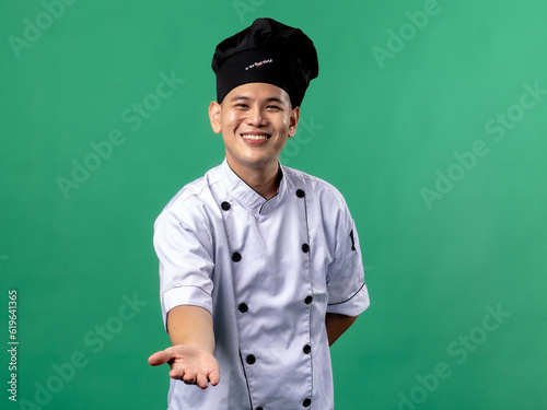 A portrait of an Indonesian Asian man wearing a chef's uniform and a chef's hat, seen posing, isolated with a green background.