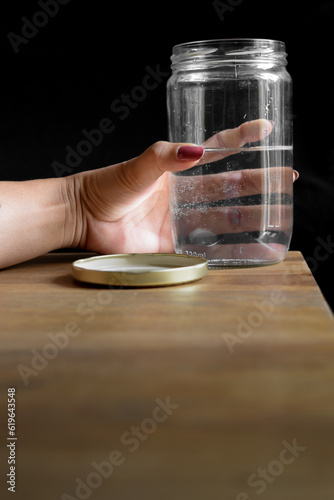 A woman's hand holds a glass jar filled with water and the jar is on a wooden table that also has a black background.