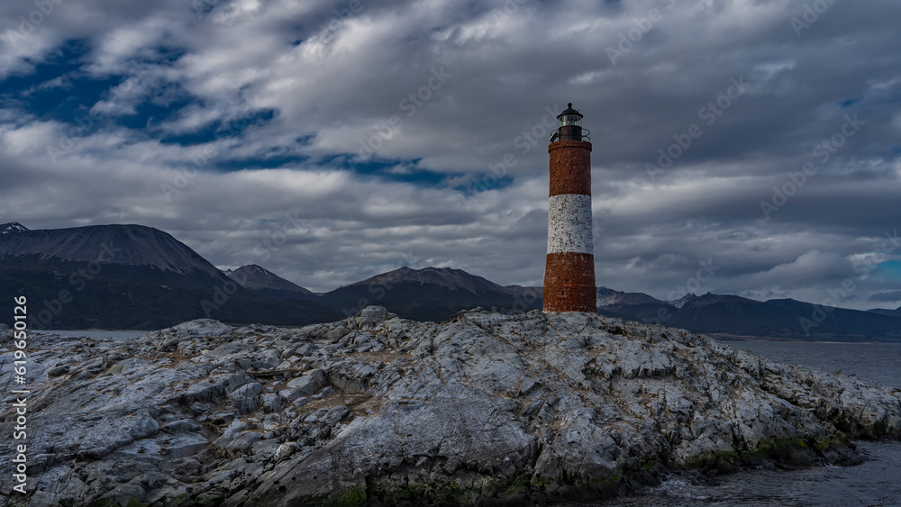 An old lighthouse Les Eclaireurs on a rocky island in the Beagle Channel. A red-and-white striped stone tower against the sky and clouds. A mountain range in the distance. Argentina.