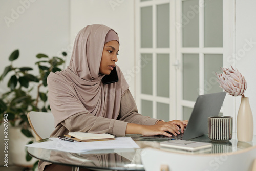 Young busy Muslim female employee with mobile phone by ear typing on laptop keyboard while organizing work by table in home environment