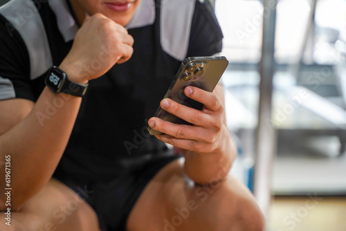 Closeup and crop Asian guy relaxing from workout with mobile phone at fitness center.