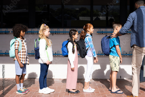 Diverse male teacher and elementary schoolchildren with schoolbags queuing in school playground