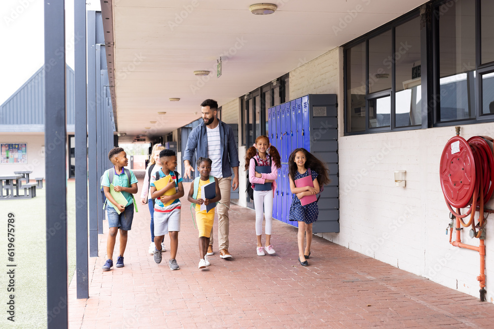 Diverse, happy male teacher and elementary schoolchildren walking in school corridor, copy space