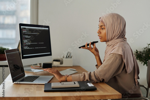 Side view of confident Muslim female programmer talking by speakerphone during work while holding mobile phone by her mouth