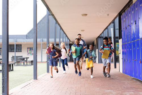 Diverse male teacher with happy children running in elementary school corridor, copy space