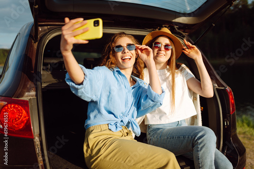 Smiling young womans have fun in the back seat of a car while traveling. Girlfriends take selfies, laugh and relax while sitting in car and enjoying the sunny weather. Active lifestyle.