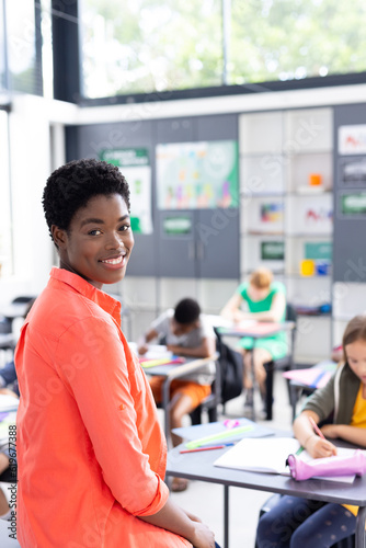 Vertical portrait of happy african american female teacher in class with diverse pupils, copy space