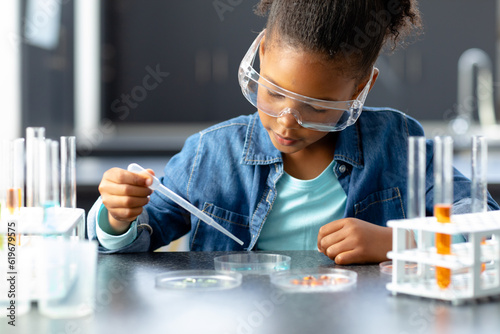 Happy biracial schoolgirl in safety glasses using pipette in science class with copy space