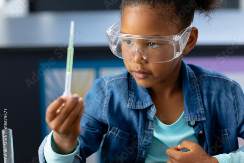 Happy biracial schoolgirl in safety glasses observing pipette in science class with copy space