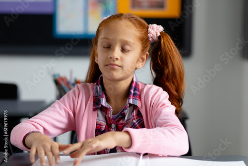 Blind, biracial schoolgirl sitting at desk reading braille in class